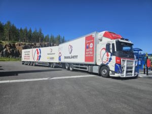 Suuri rekka, jossa on pitkä perävaunu, pysähtyneenä kauniissa maisemassa. Taustalla näkyy metsää ja kirkas sininen taivas. Rekassa on yrityksen logoja ja mainoksia, jotka korostavat sen kuljetuspalveluja.