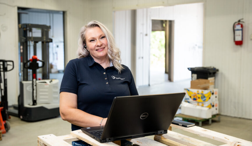 Nainen hymyilee tietokoneen äärellä teollisuusympäristössä, jossa näkyy varastokoneita ja pakkausmateriaaleja taustalla. Hänellä on päällään tummansininen polo-paita ja hän käyttää kuulokkeita. Kuvassa korostuu moderni työympäristö ja etätyön mahdollisuudet teollisuusalalla.